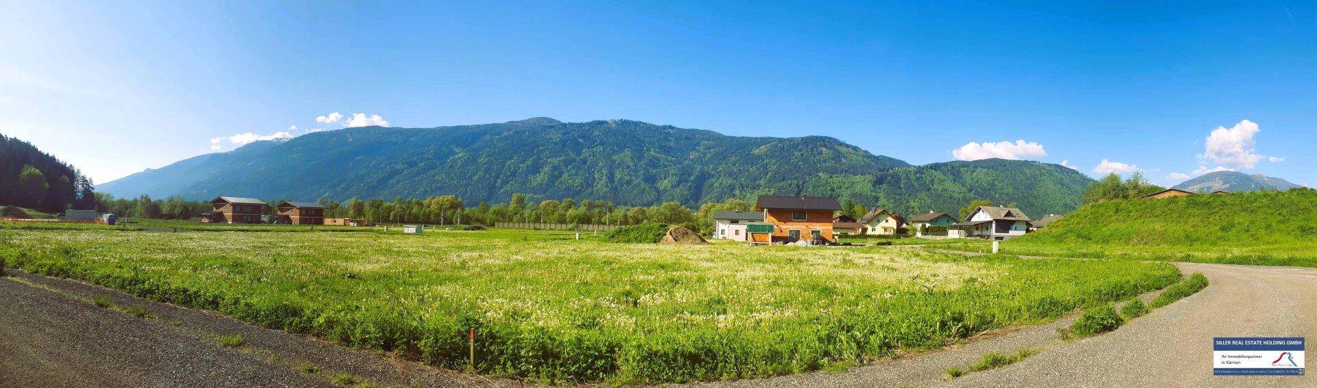 Panoramablick auf eine ländliche Siedlung mit Häusern, grünen Feldern und einer beeindruckenden Bergkette unter klarem Himmel.