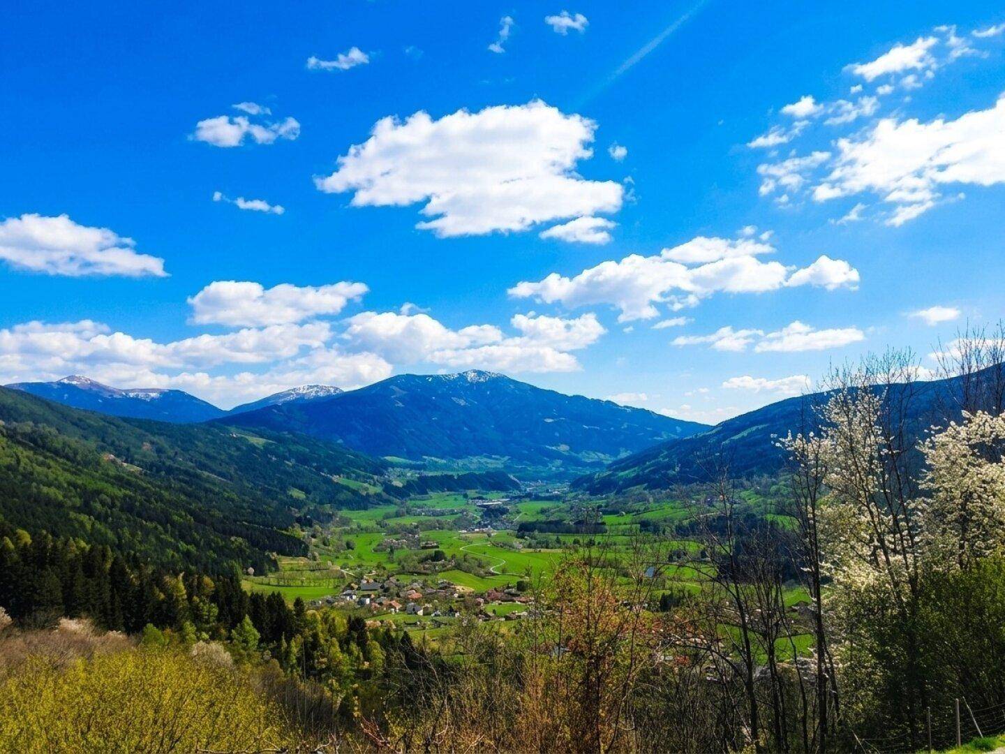 Malerisches Tal mit einem kleinen Dorf, umgeben von grünen Wäldern und schneebedeckten Bergen.