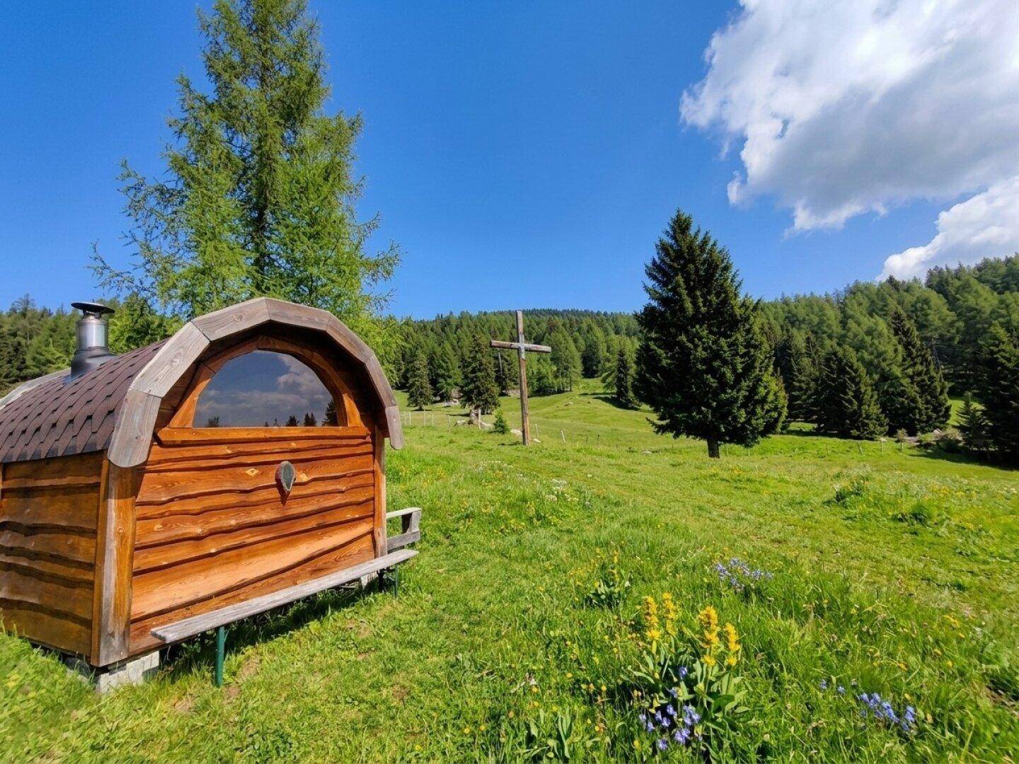 Rustikale Holzfass-Sauna auf einer grünen Wiese mit einem großen Holzkreuz und Wald im Hintergrund.