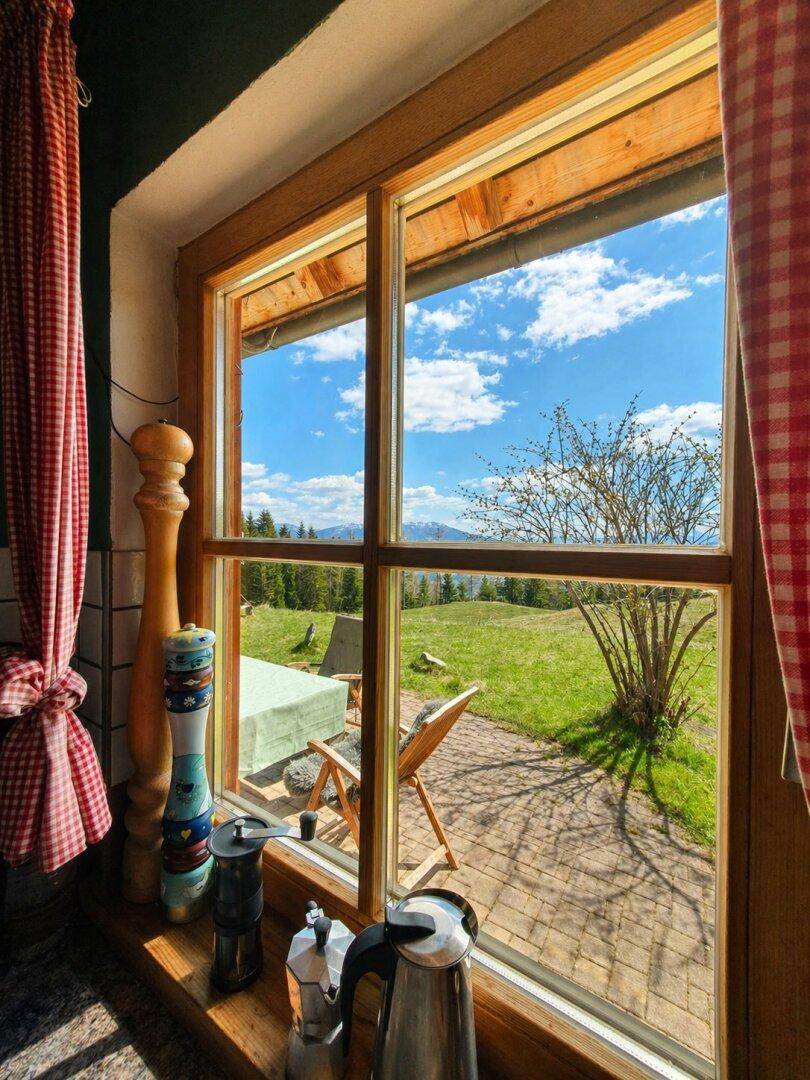 Blick aus einem Holzfenster auf eine gepflasterte Terrasse und die weite grüne Landschaft mit Bergen.