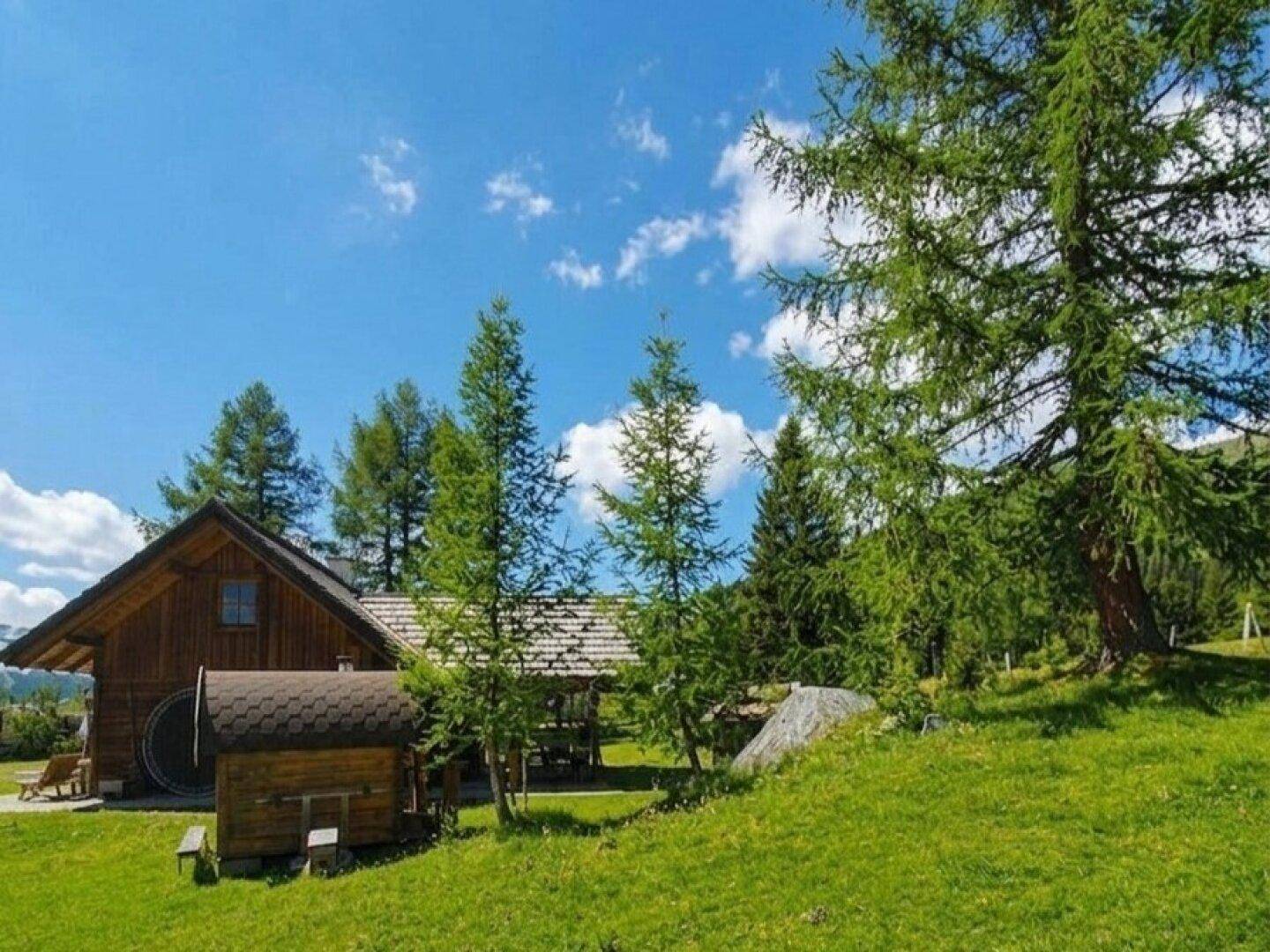 Idyllisches Holzhaus mit einer kleinen Holzfass-Sauna, umgeben von hohen Bäumen und grünen Wiesen.
