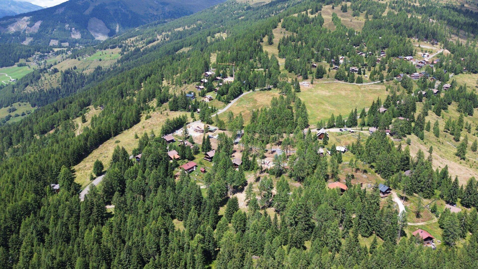 Luftaufnahme eines Almdorfes inmitten einer weitläufigen Berglandschaft mit dichten Wäldern.