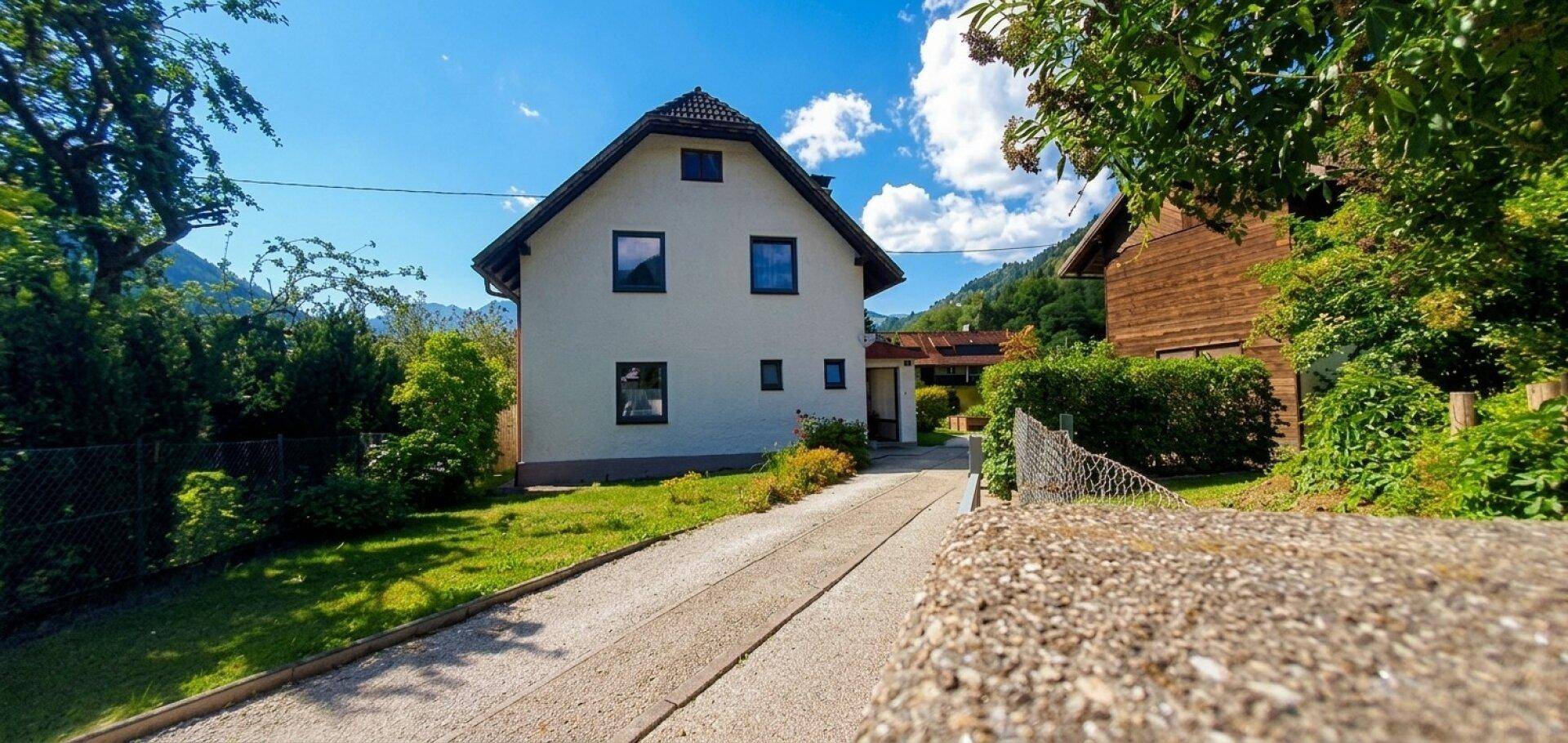 Vorderansicht des traditionellen weißen Hauses mit Satteldach, Auffahrt und grüner Umgebung.