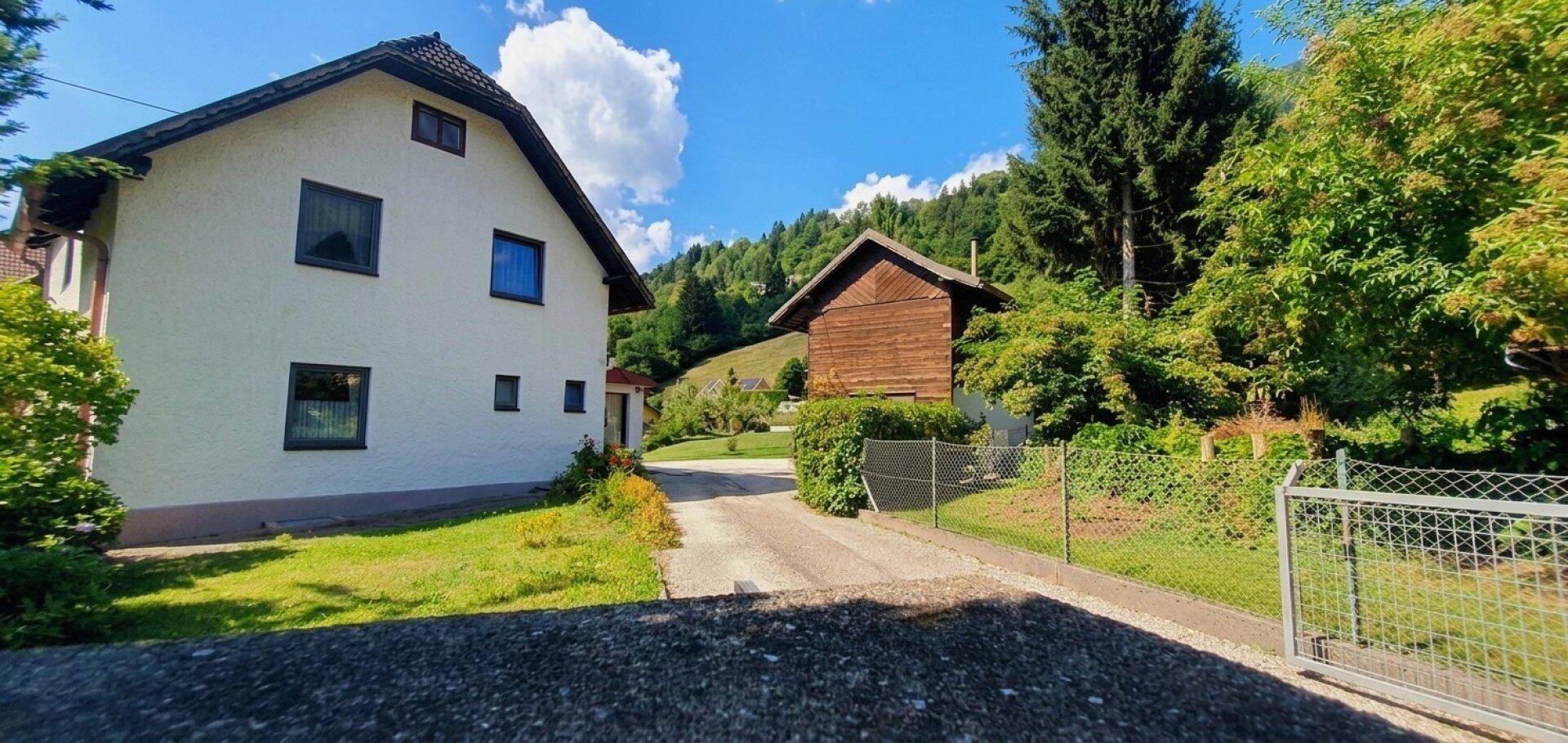 Ansicht des Haupthauses und des Holzgebäudes mit Auffahrt und Zaun an einem sonnigen Tag.