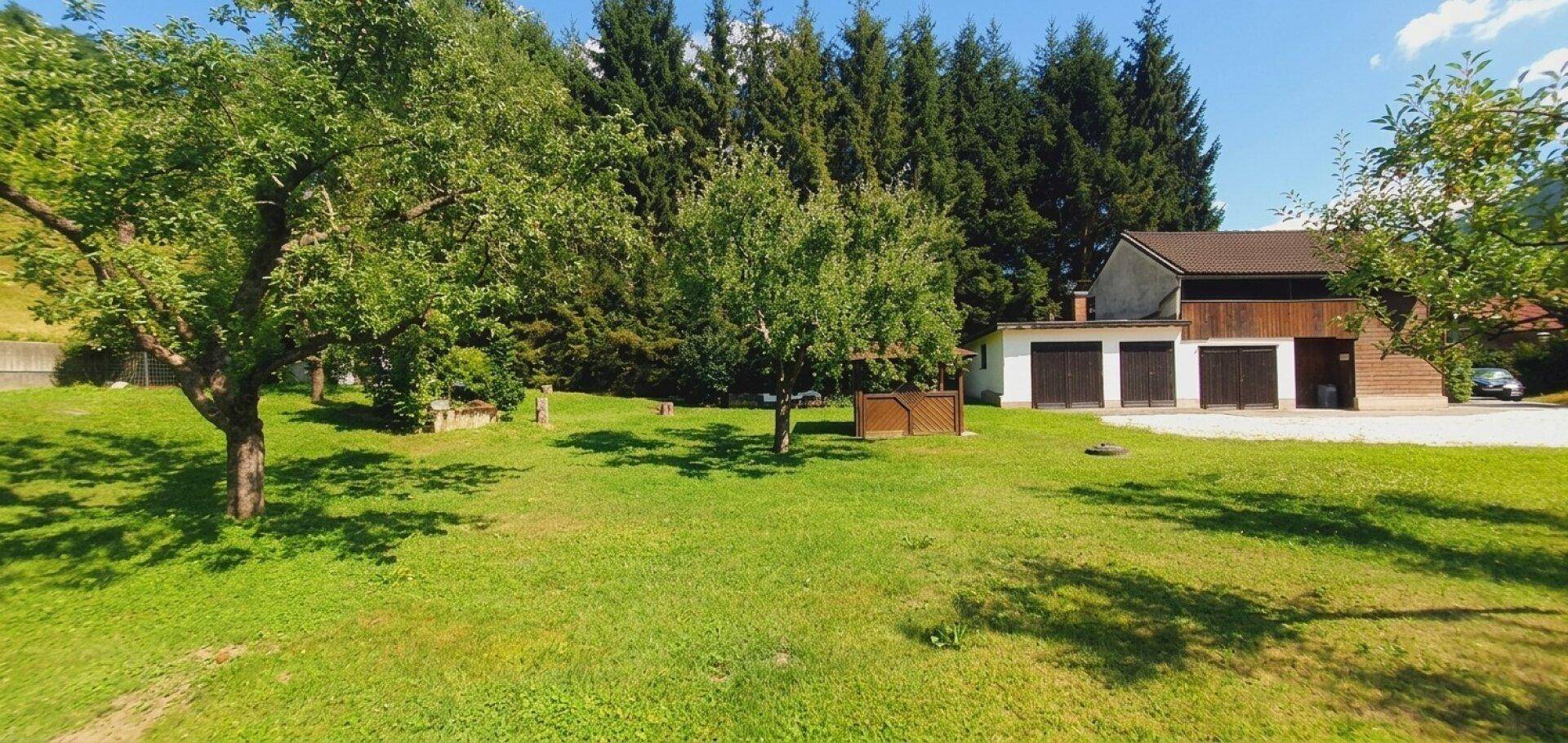 Großer Gartenbereich mit Rasen und Bäumen, der zu einem Gebäude mit Garagen führt, umgeben von Wald.