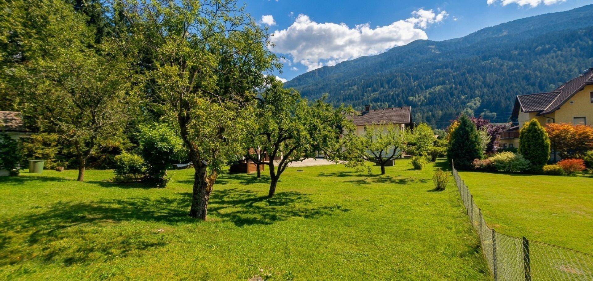 Weitläufiger Garten mit grüner Rasenfläche, alten Bäumen und einem Zaun, umgeben von Bergen.