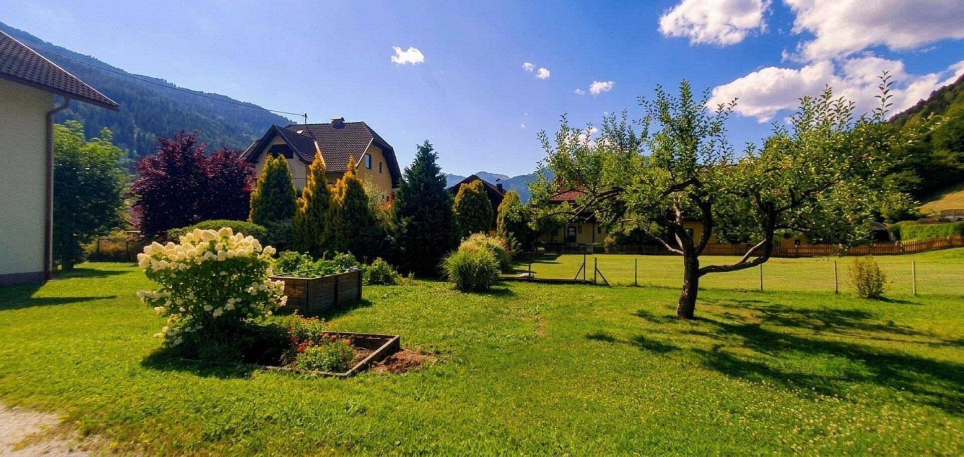 Weitläufiger Garten mit Rasen, Bäumen, Hochbeet und einem Haus im Hintergrund vor Bergkulisse.