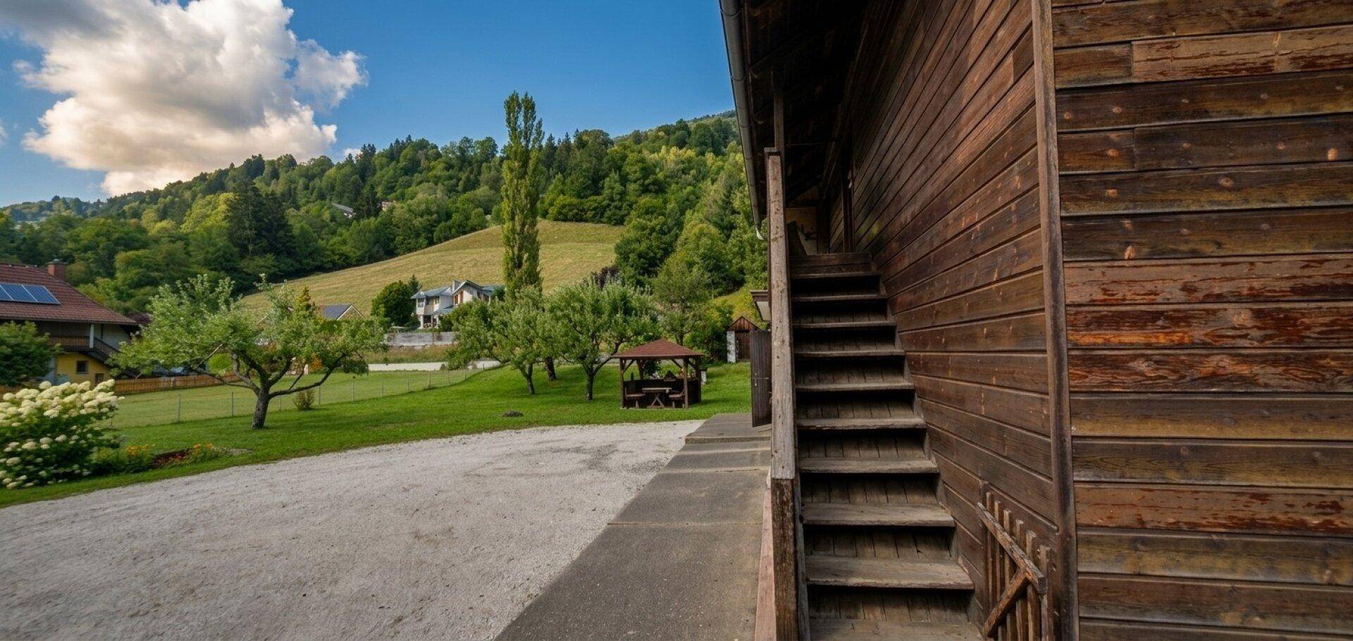 Seitenansicht des Holzgebäudes mit Außentreppe und Kiesweg, umgeben von Garten und Bergen.