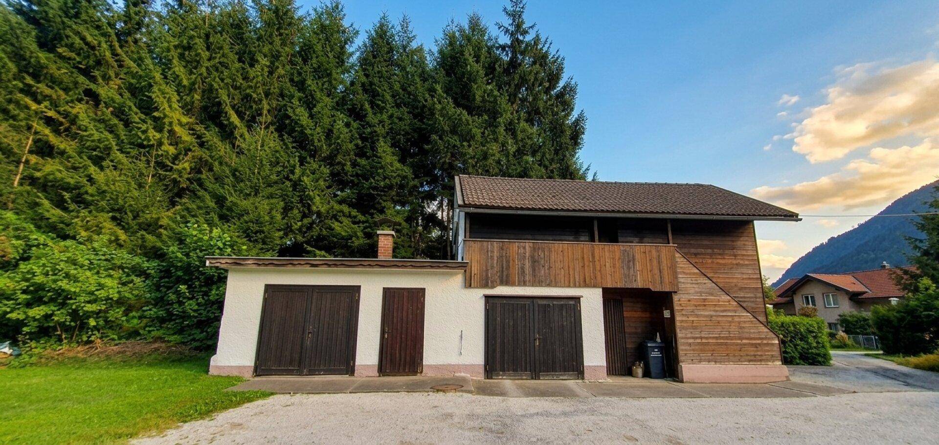 Rustikales Nebengebäude mit zwei Garagentoren und Holzverkleidung, umgeben von dichtem Wald.