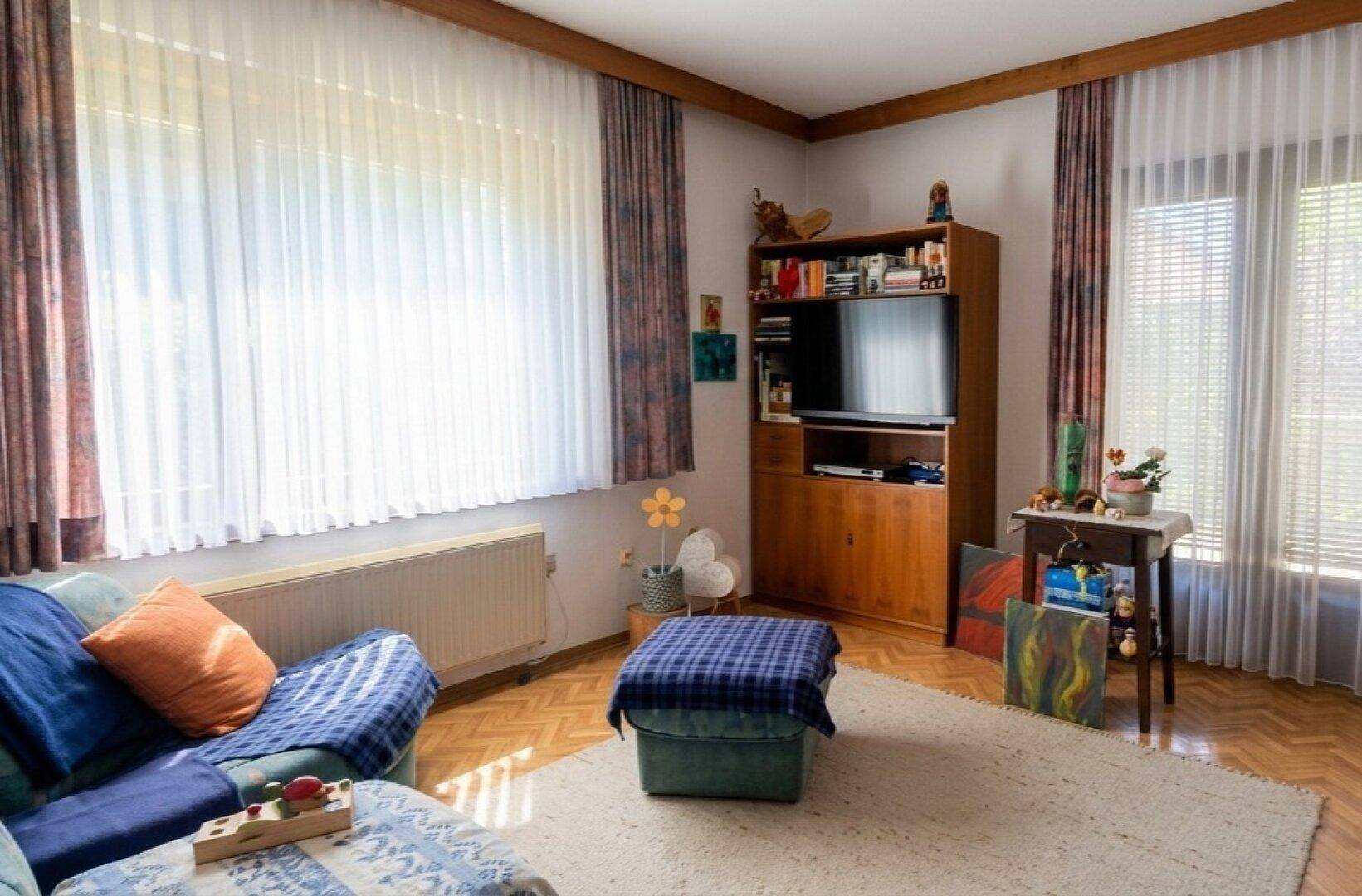 Helles Wohnzimmer mit blauem Sofa, Holzmöbeln, Fernseher und großem Fenster mit Parkettboden.