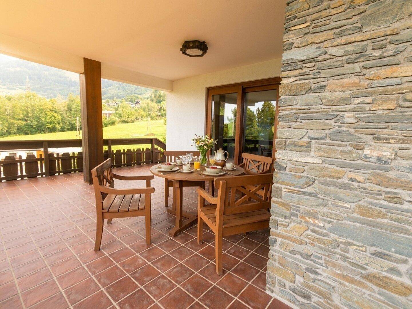 Überdachte Terrasse mit Esstisch und Stühlen, umgeben von Natur und Bergblick.