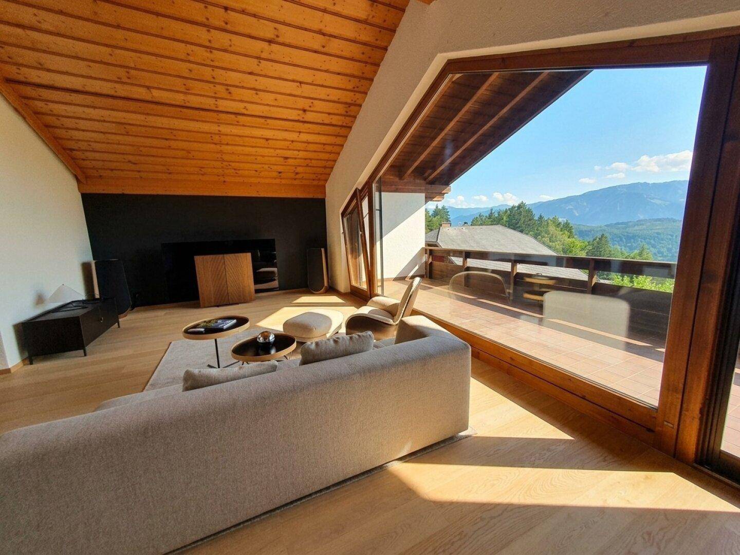 Lichtdurchflutetes Wohnzimmer mit Holzbalkendecke, großen Fenstern und Panoramablick auf die Berge.