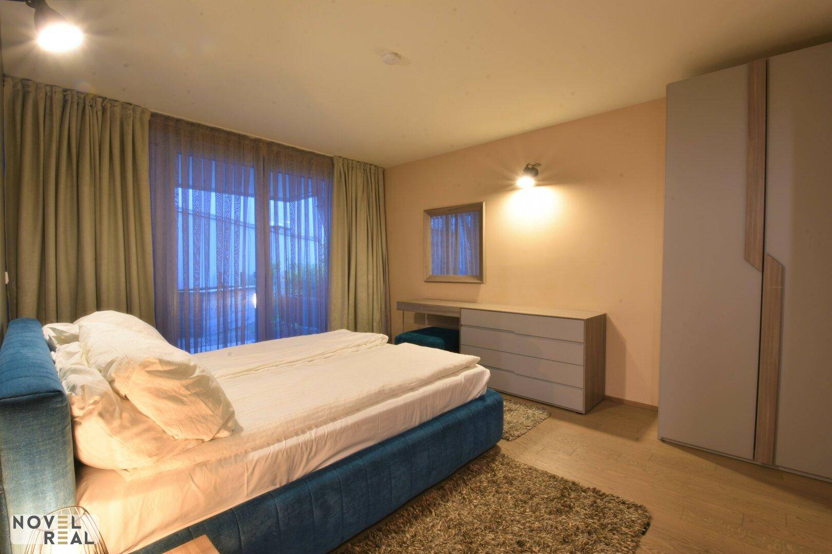 Gemütliches Schlafzimmer mit blauem Bett, großem Kleiderschrank und Vorhängen an den Fenstern.
