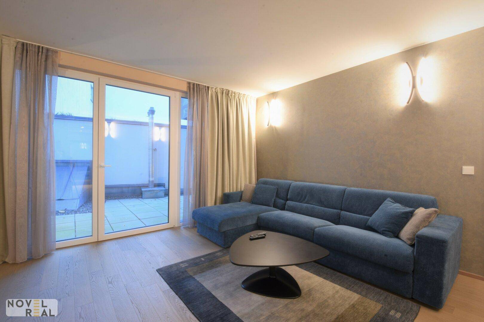 Helles Wohnzimmer mit bequemer blauer Couch und großem Fenster zur Außenterrasse.