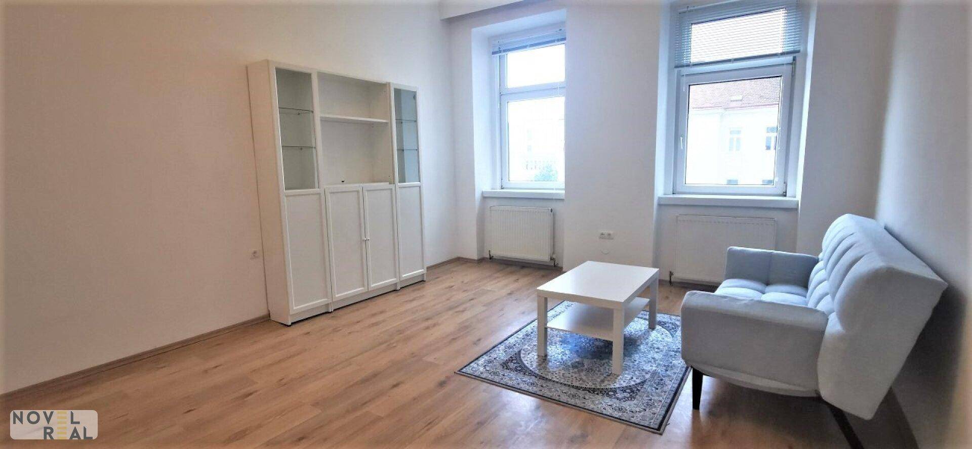 Geräumiges Wohnzimmer mit hellem Laminatboden, Sofa und weißem Regal.