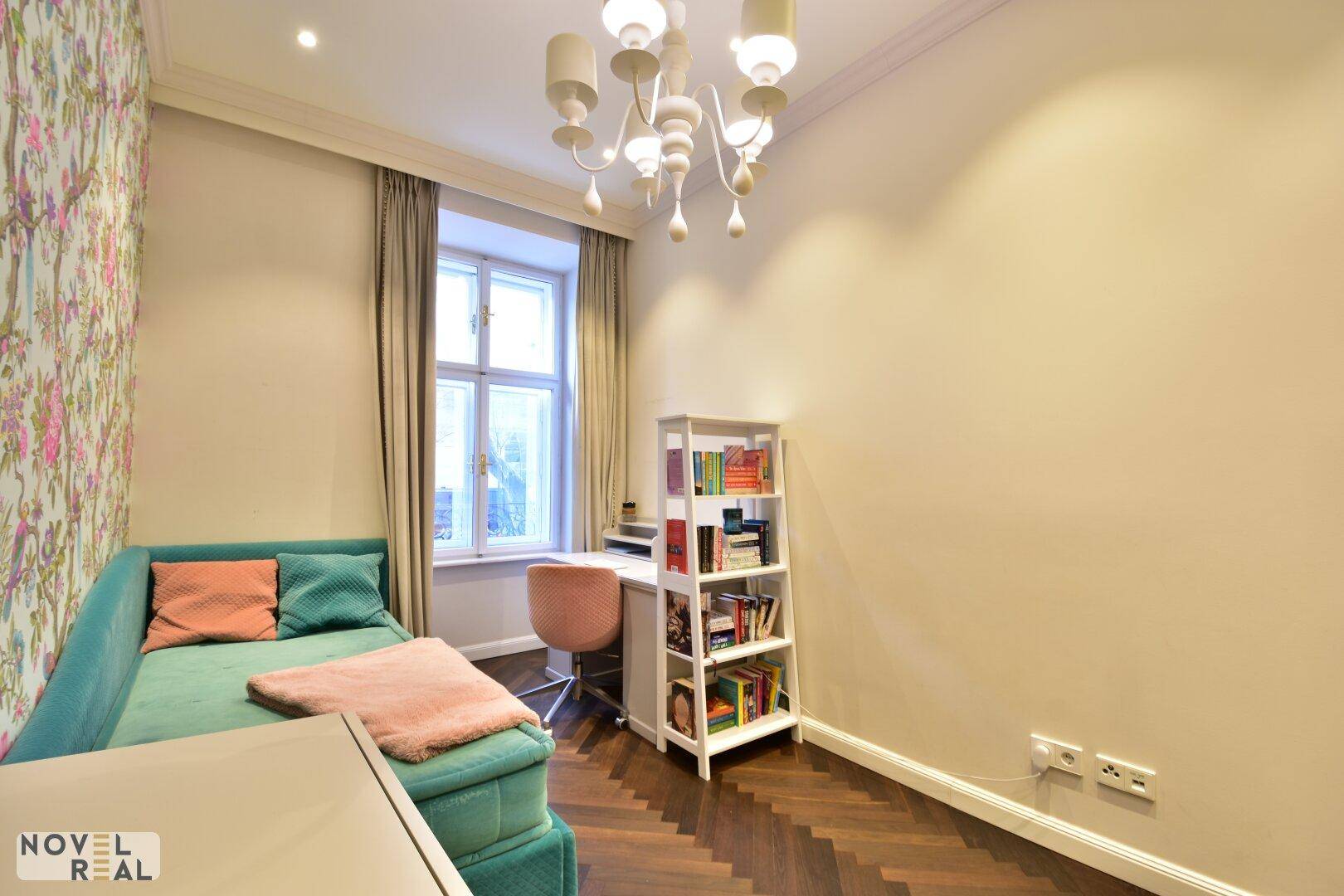 Helles Schlafzimmer mit floraler Tapete, Parkettboden und einem Schreibtisch am Fenster.