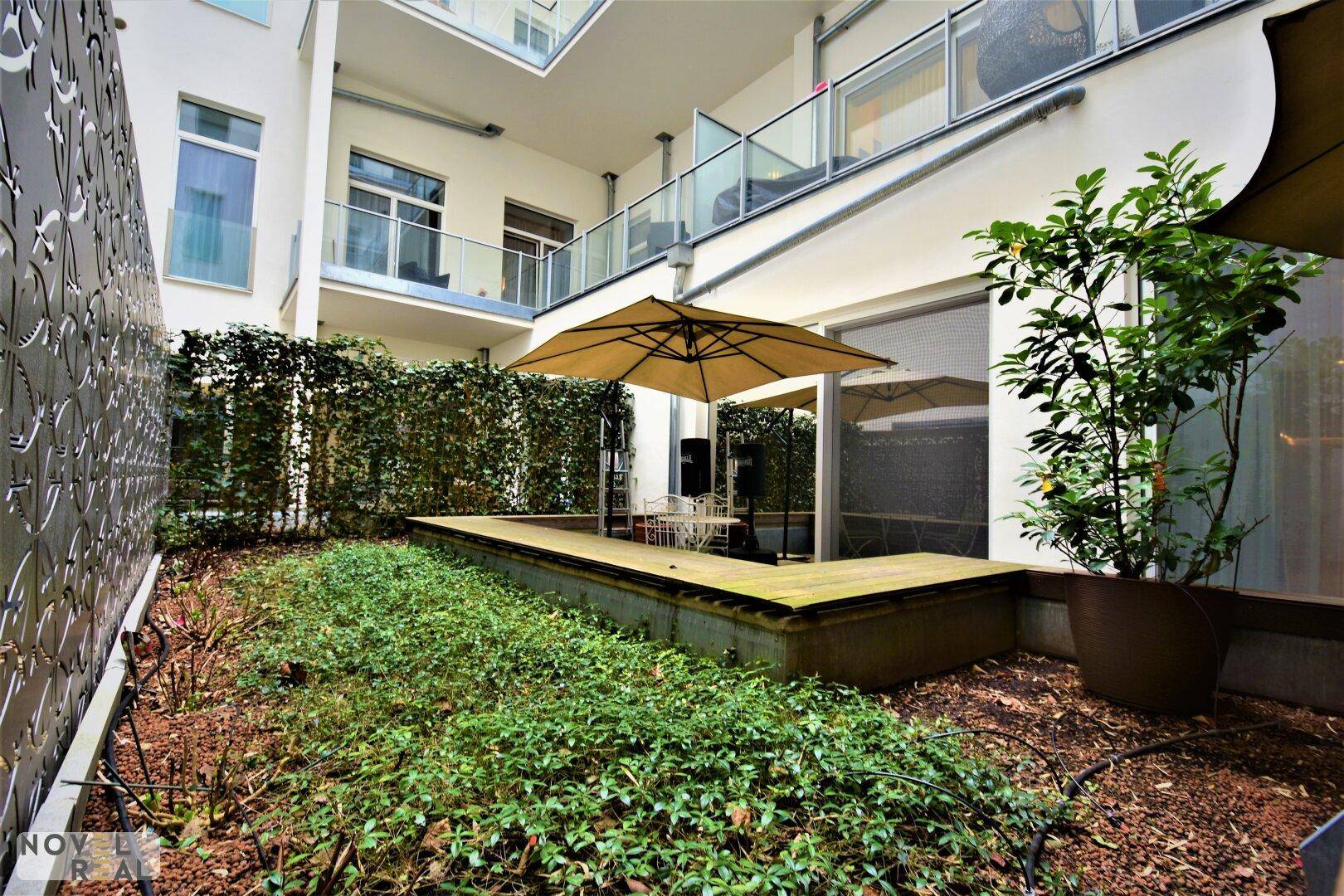 Grüner Innenhof mit kleiner Terrasse, umgeben von Hecken und Gebäudefassaden.