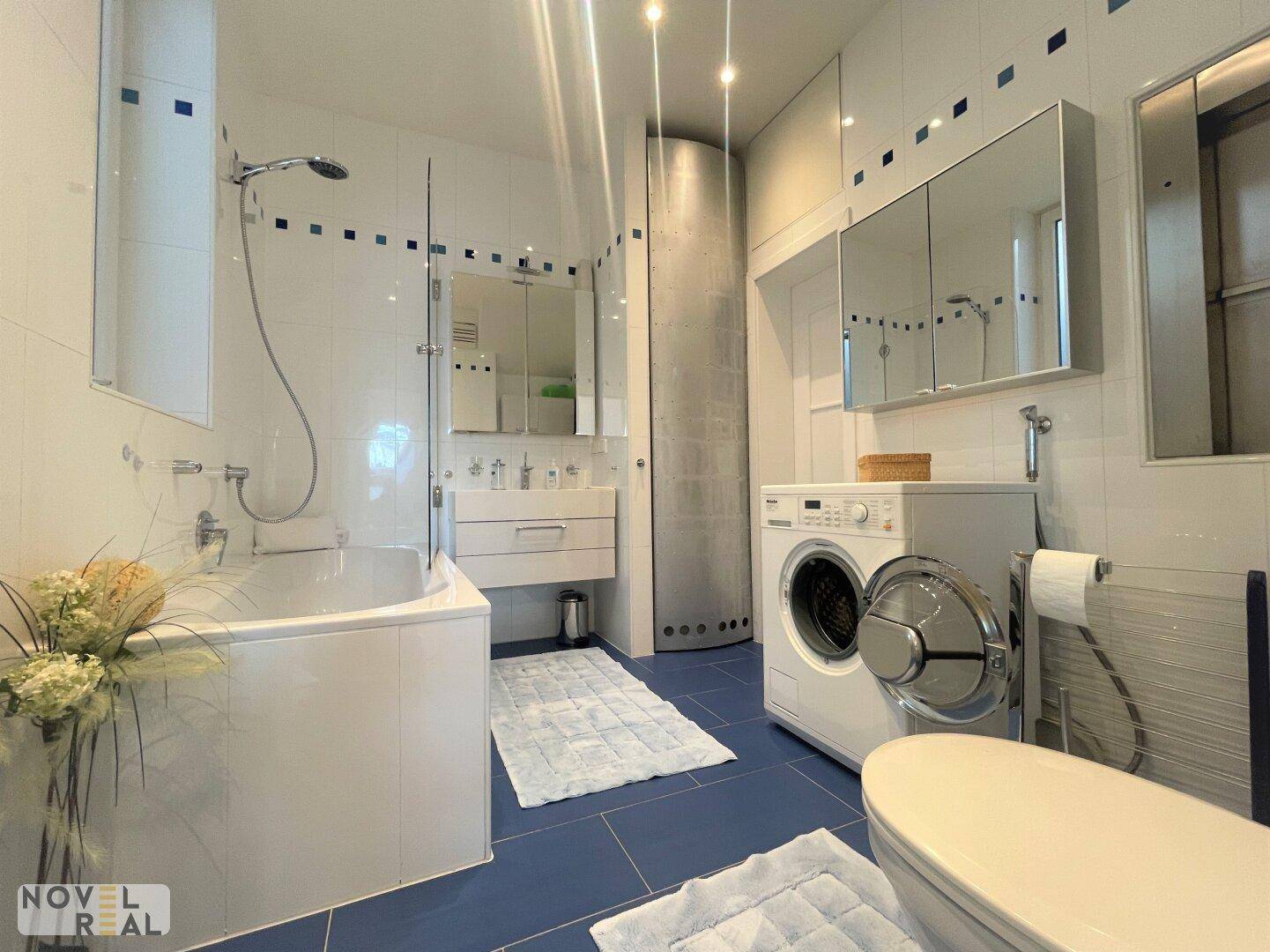 Badezimmer mit Badewanne, Dusche, Waschmaschine und blauen Bodenfliesen.