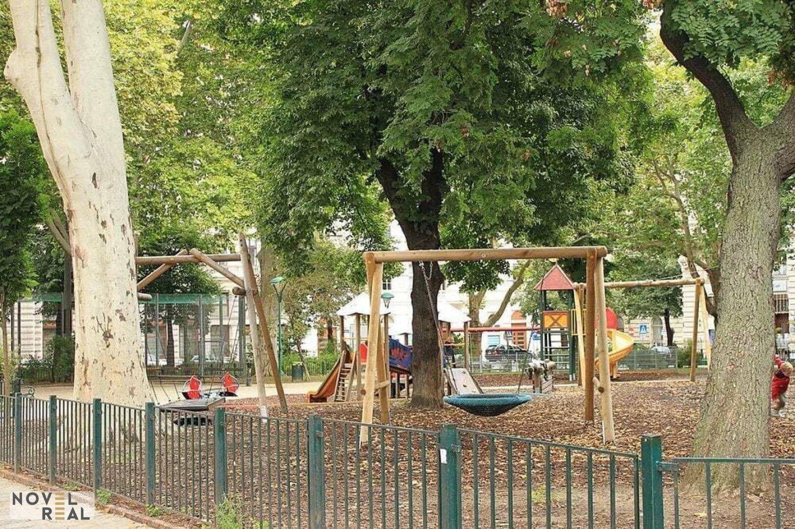 Grüner Spielplatz mit Schaukeln und Rutschen, umgeben von Bäumen und einem Zaun.