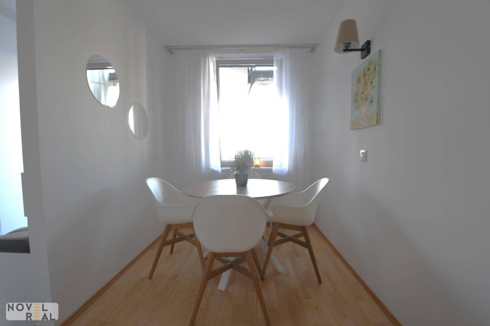 Heller Essbereich mit rundem Holztisch und modernen weißen Stühlen am Fenster.