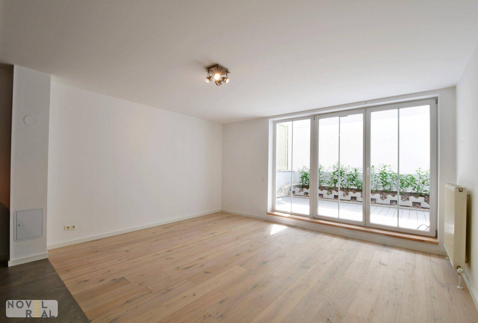 Heller, leerer Raum mit Holzboden und großen Fenstern, die auf eine Terrasse blicken.