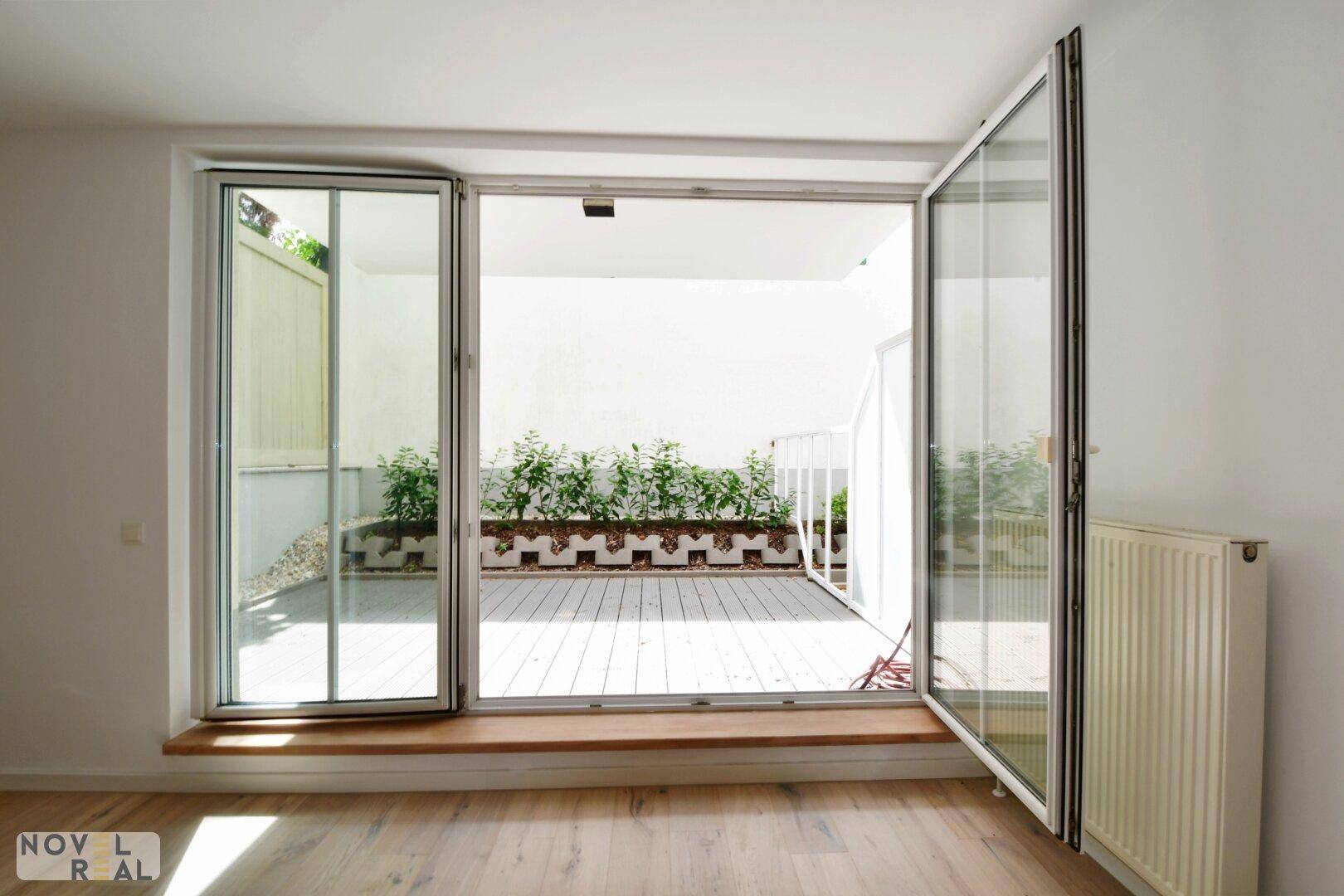 Heller Raum mit geöffneten Türen, die auf eine begrünte Terrasse führen.