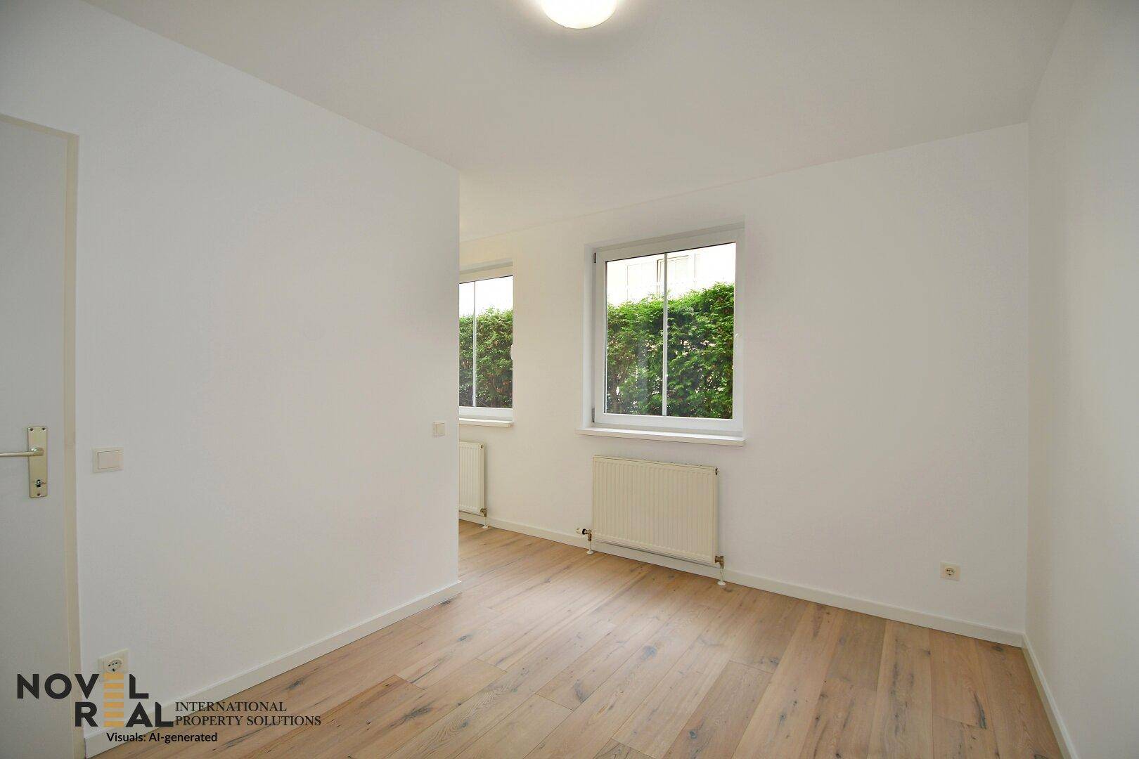 Helles Zimmer mit zwei Fenstern, Holzboden und weiß gestrichenen Wänden.