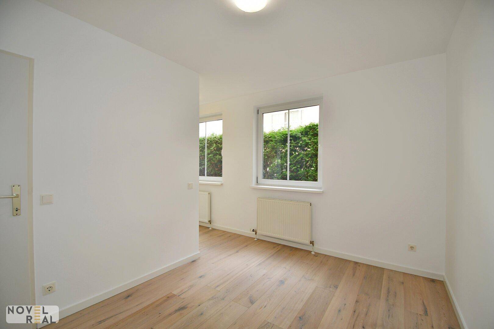 Helles Zimmer mit Holzboden und zwei Fenstern, die einen grünen Ausblick bieten.