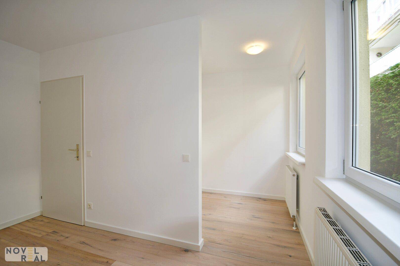 Heller, leerer Raum mit Holzboden und einem Fenster, das Tageslicht hereinlässt.