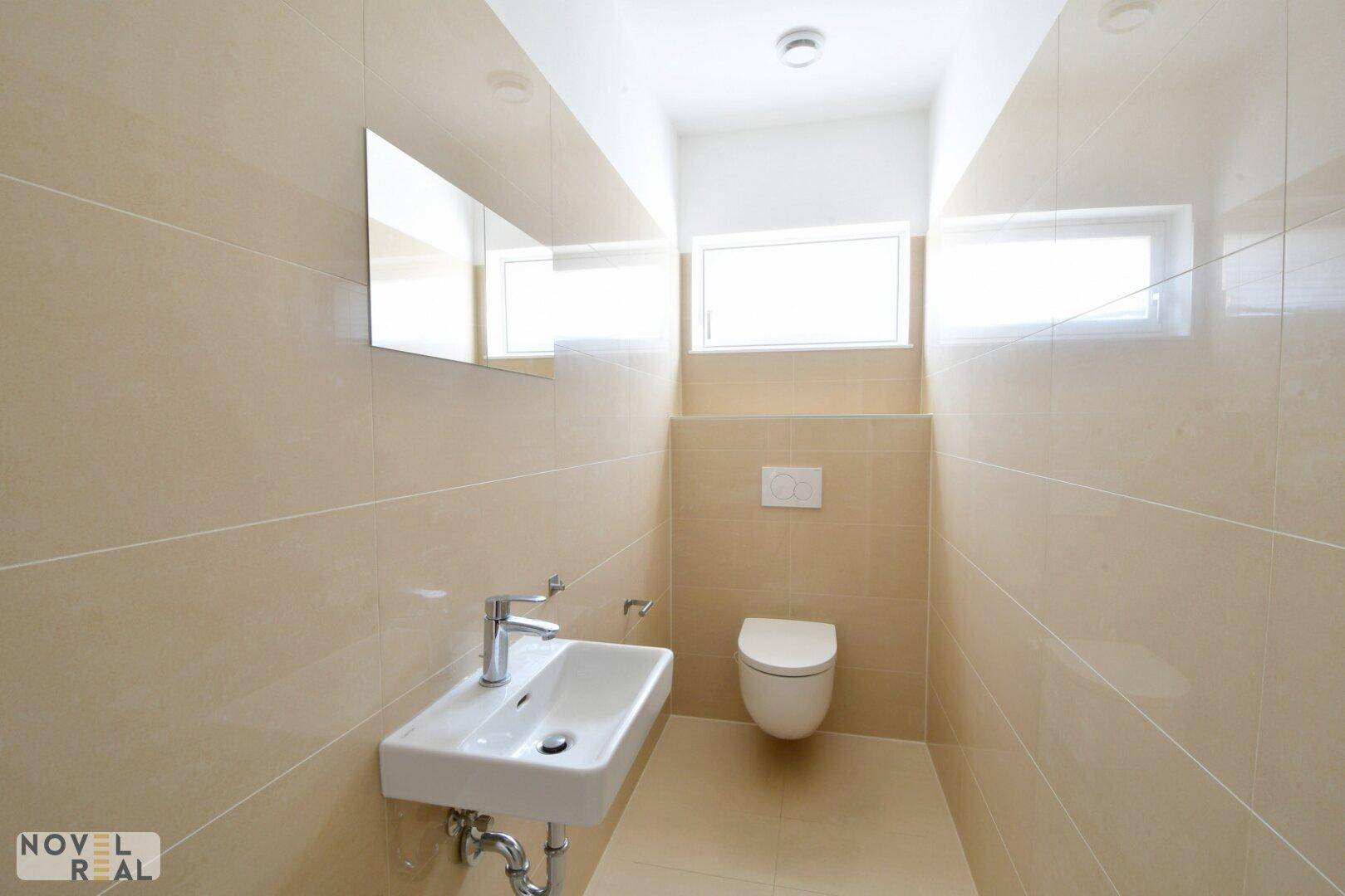 Modernes WC mit beigen Fliesen, wandhängendem WC und kleinem Waschbecken.