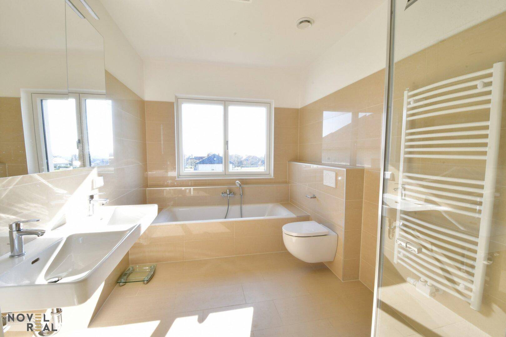 Modernes Badezimmer mit Badewanne, Doppelwaschbecken, beigen Fliesen und Fenster.
