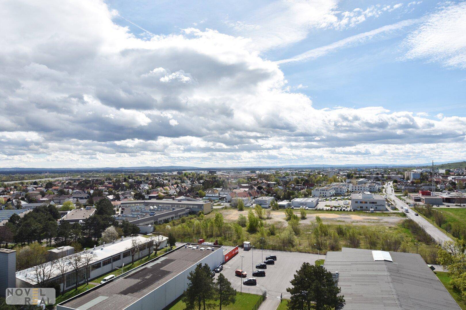 Weitläufige Luftaufnahme einer Stadt mit Gebäuden, Grünflächen und einer Straße unter bewölktem Himmel.