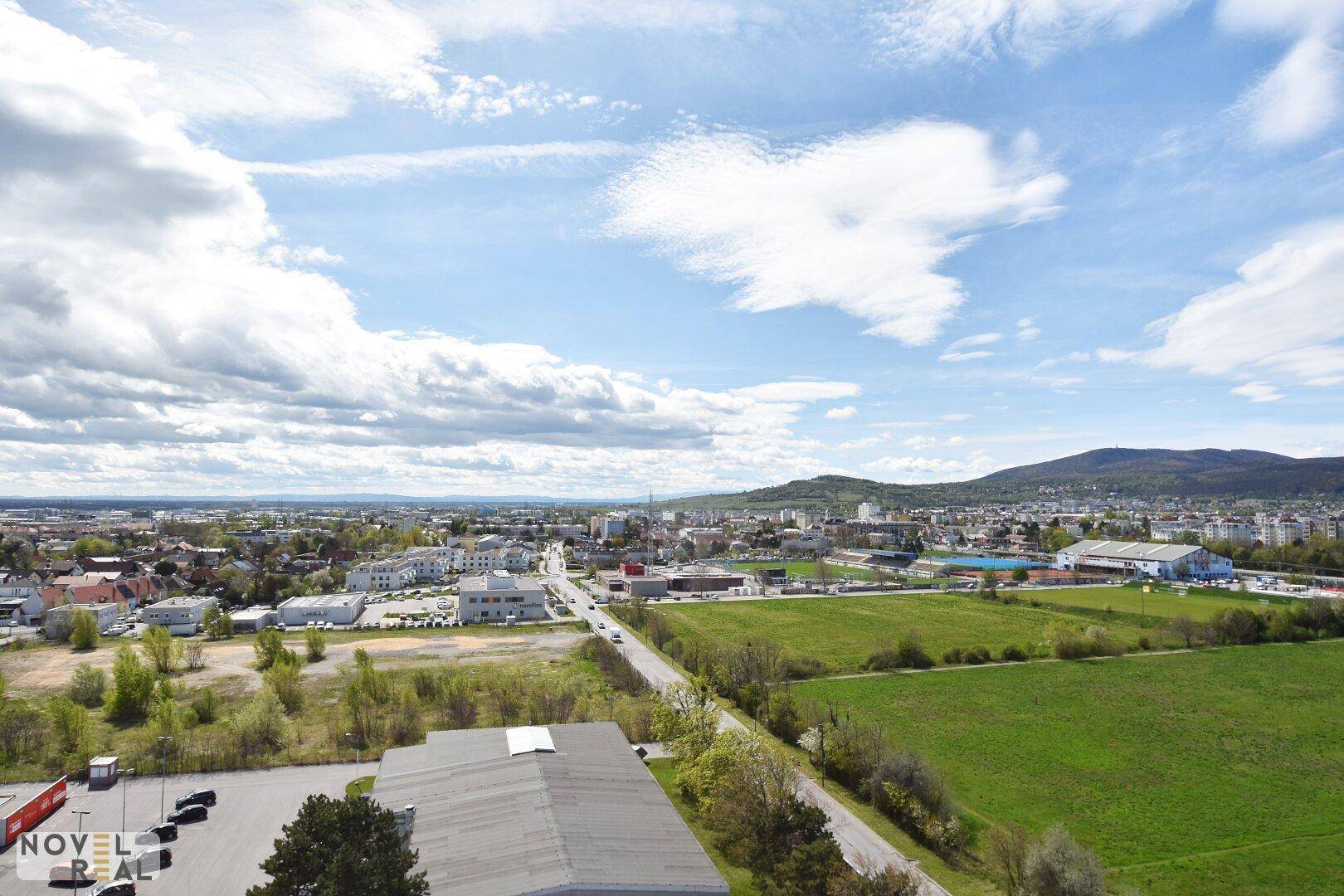 Panoramablick über eine Stadt mit Feldern, Gebäuden und Bergen im Hintergrund unter blauem Himmel.