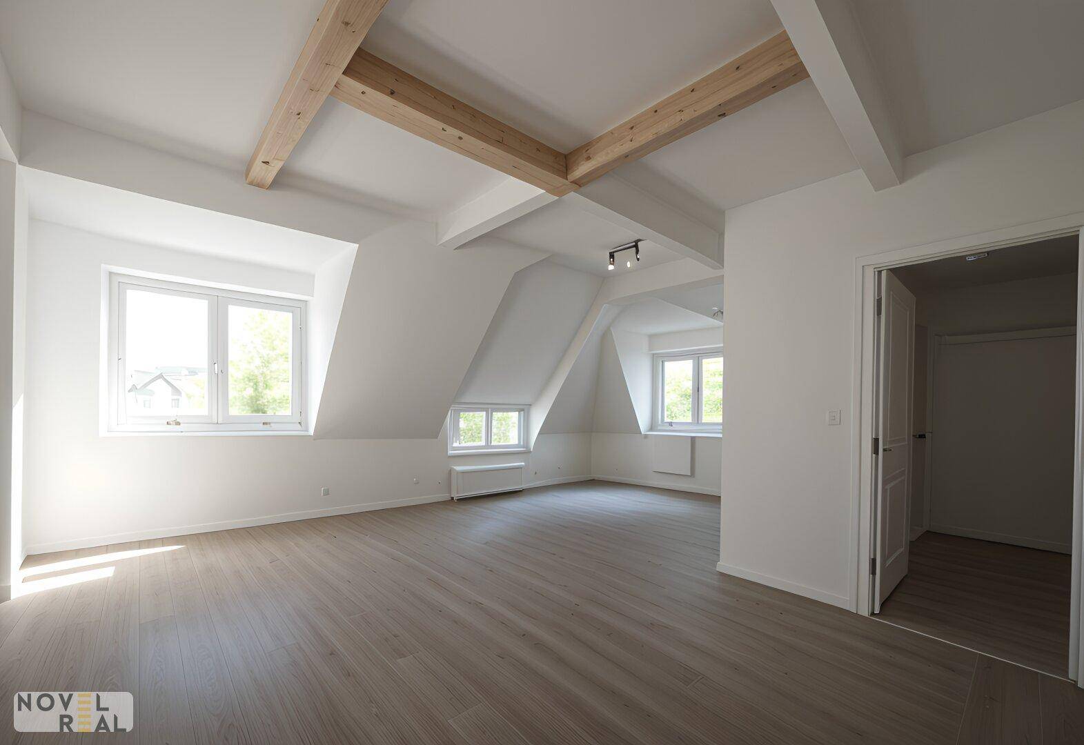 Geräumiger, heller Raum mit schrägen Wänden, sichtbaren Holzbalken und mehreren Fenstern.