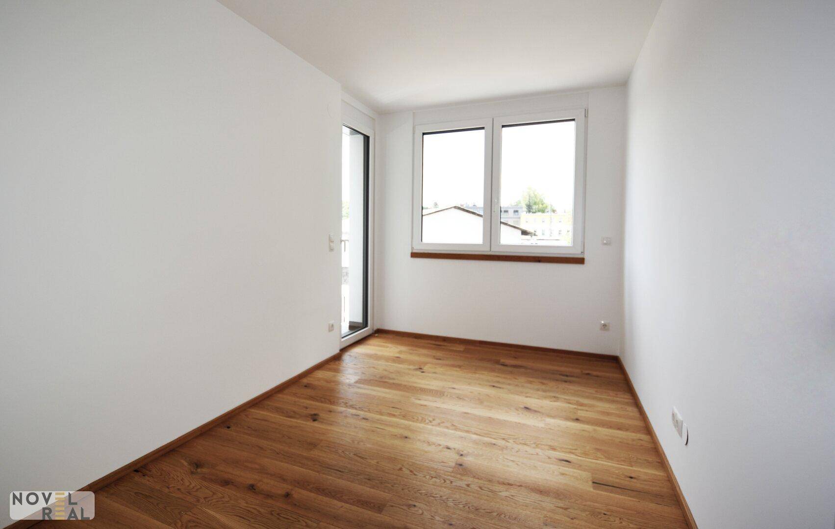 Leeres Zimmer mit Parkettboden und Zugang zu einem Balkon, bietet viel natürliches Licht.