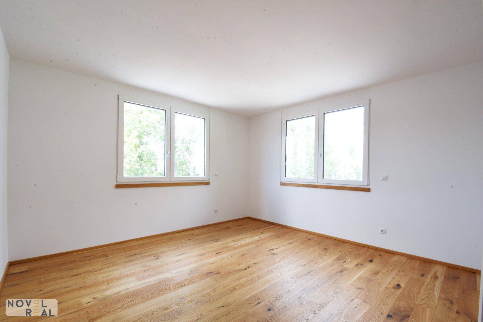 Leeres Schlafzimmer mit hellem Parkettboden und zwei großen Fenstern, die viel Tageslicht hereinlassen.