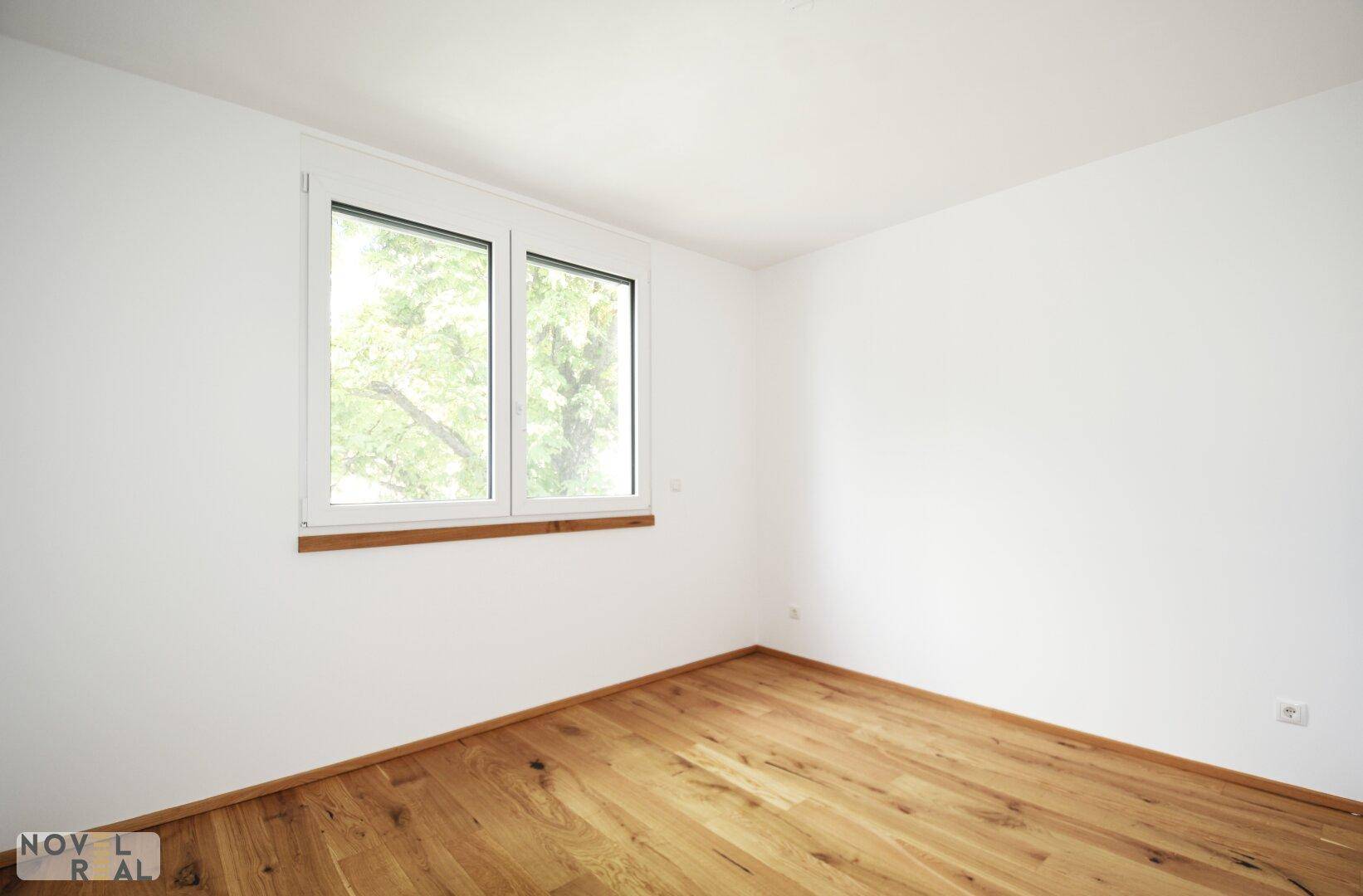 Helles Zimmer mit Holzboden und großem Fenster, das einen Blick ins Grüne bietet.