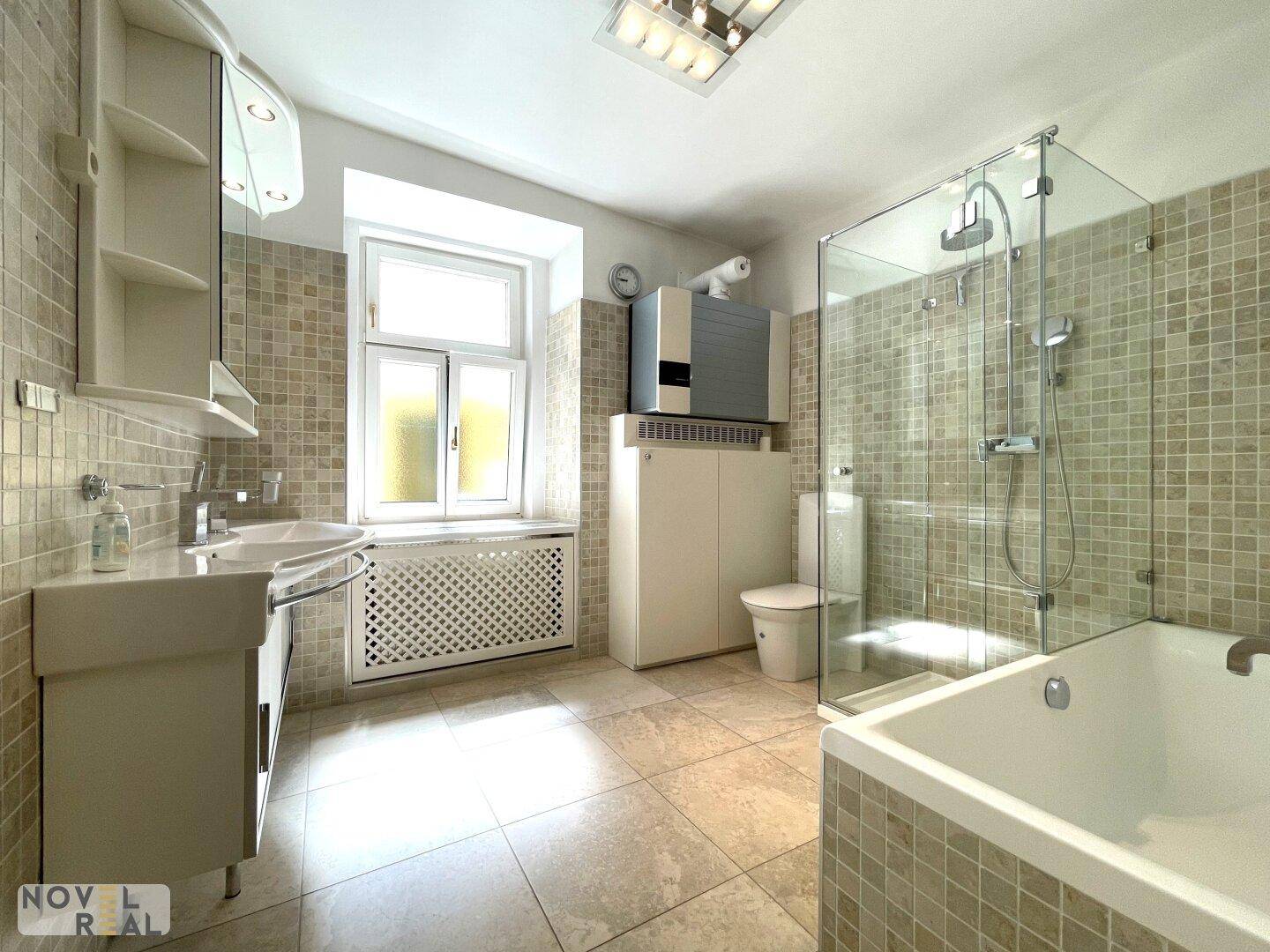 Helles Badezimmer mit Badewanne, Dusche, Fenster und beigen Fliesen.