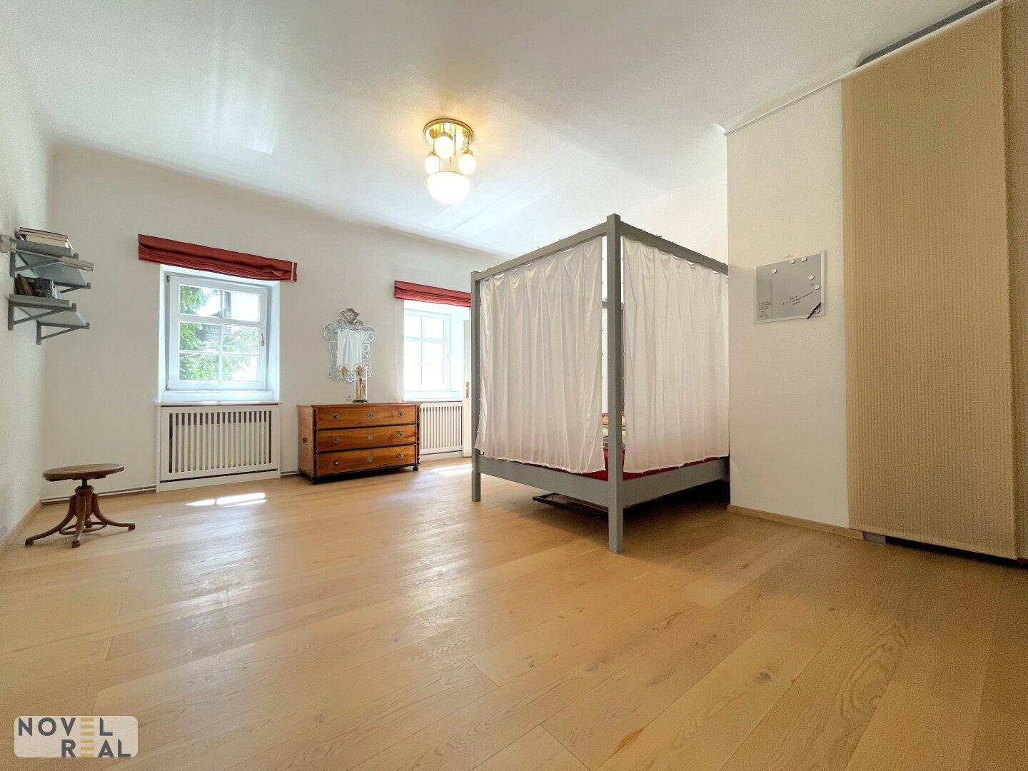 Großes Schlafzimmer mit Holzboden, Himmelbett und zwei Fenstern.
