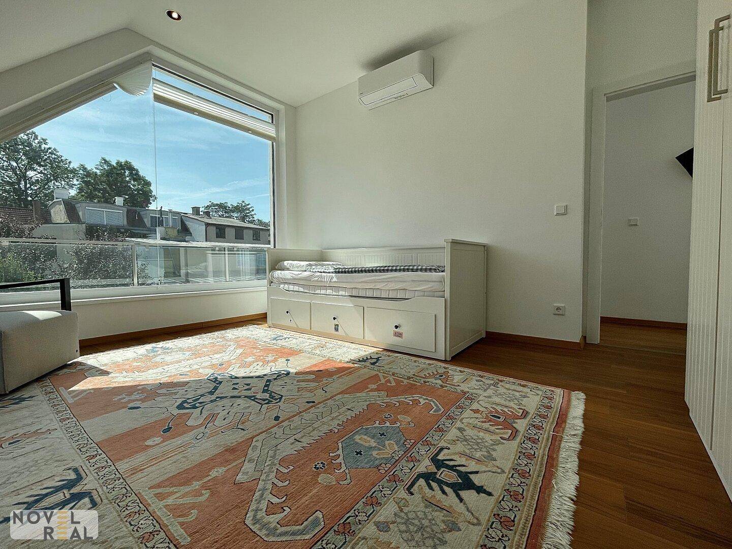 Helles Schlafzimmer mit Tagesbett, großem Fenster, Klimaanlage und Holzboden.