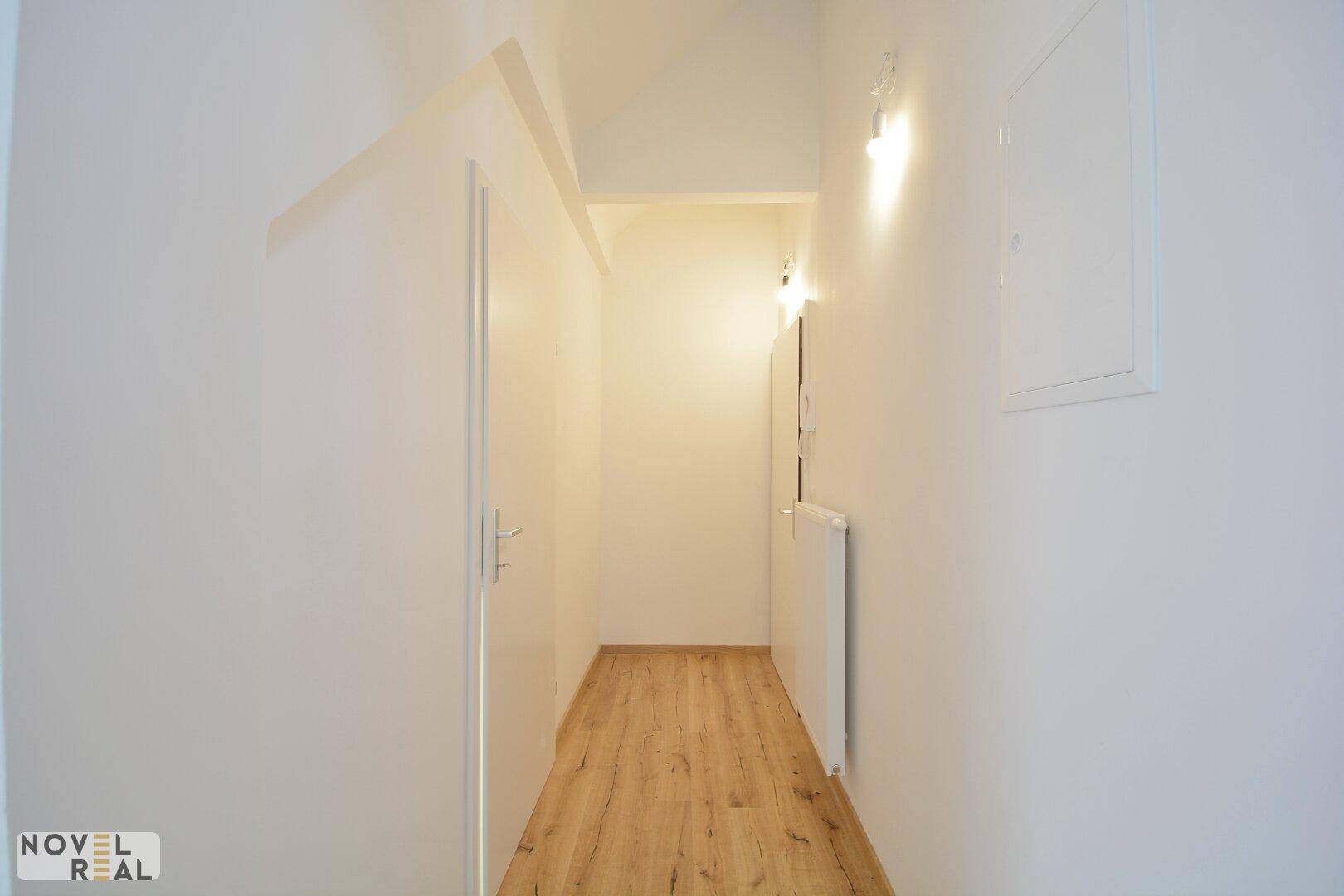 Schmaler, heller Flur mit Laminatboden und weißen Wänden, beleuchtet von Wandlampen.