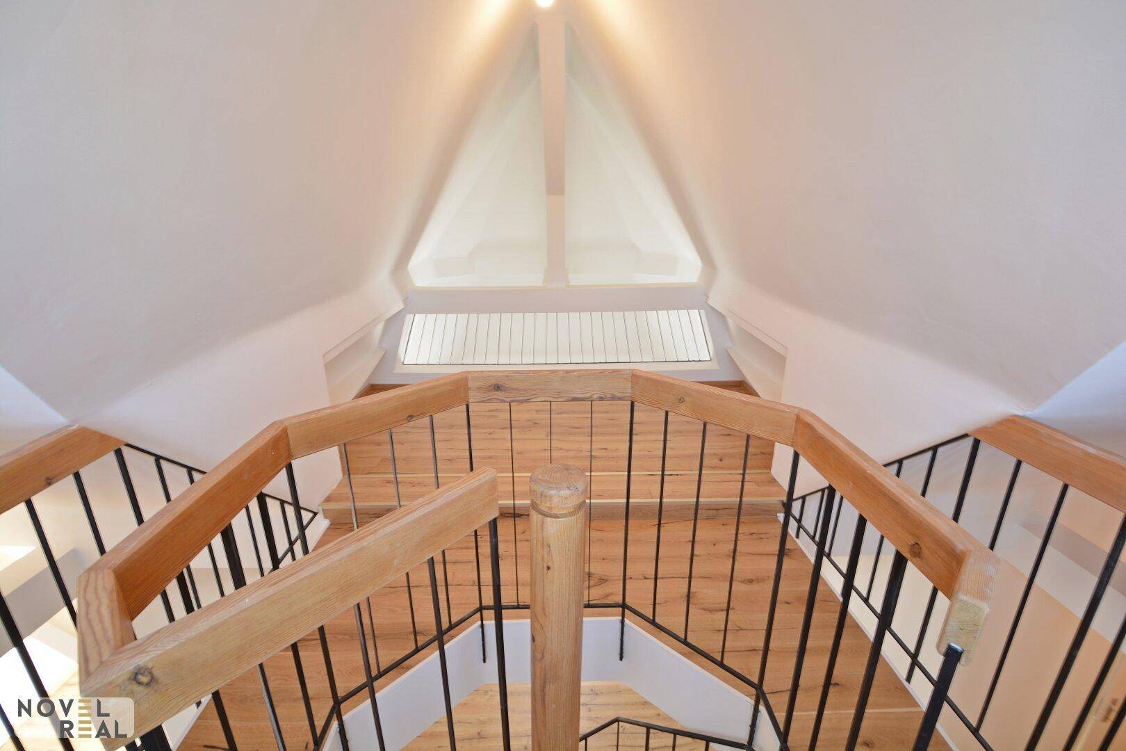 Aufsicht auf eine Holz-Wendeltreppe mit schwarzem Geländer und einem Dachfenster im oberen Bereich.