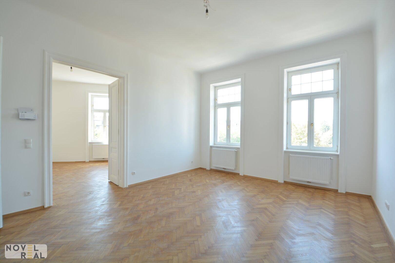 Lichtdurchfluteter Raum mit Fischgrätparkett und hohen Fenstern, ideal für ein Wohnzimmer.