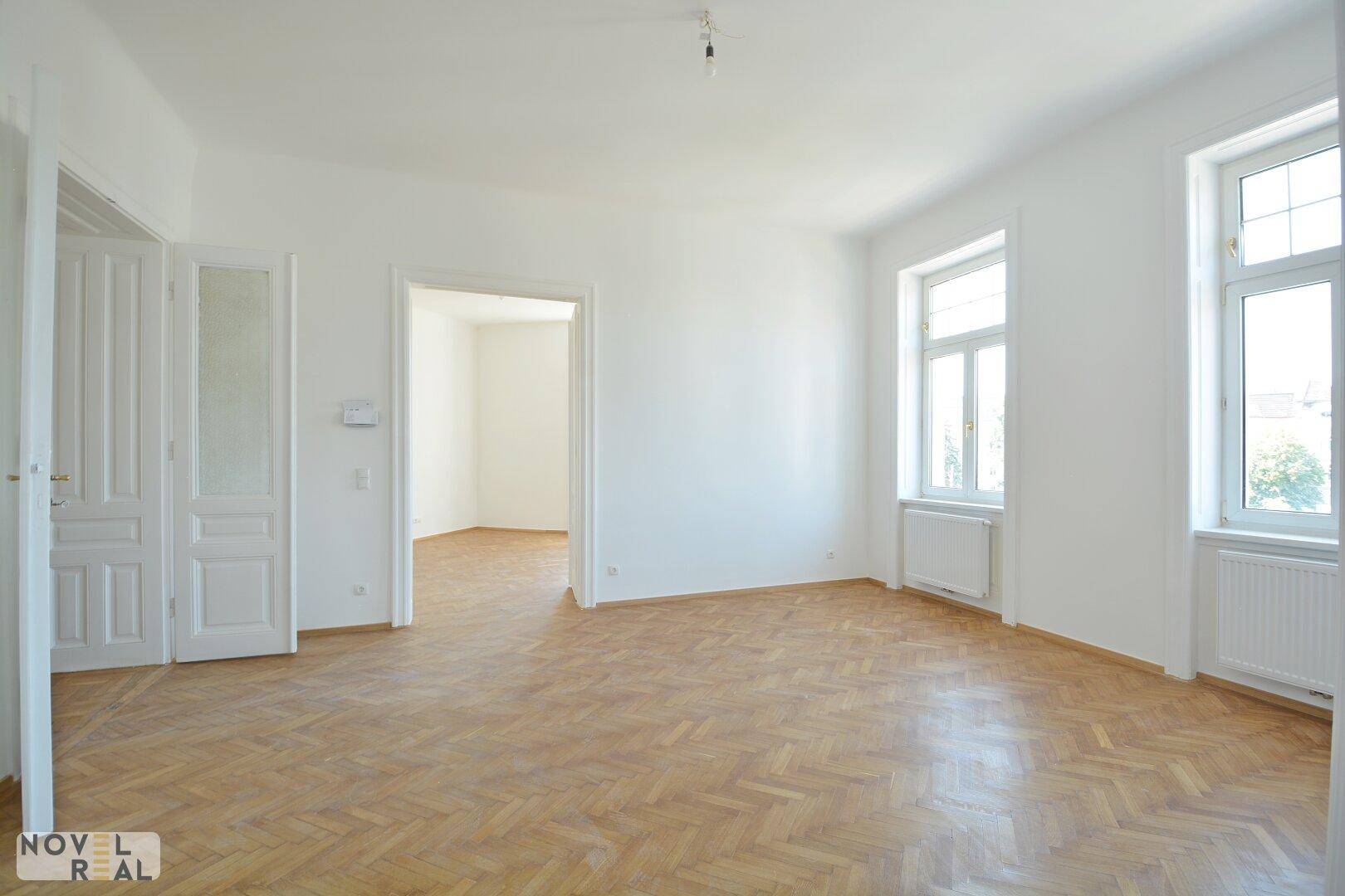 Heller Raum mit Fischgrätparkett und hohen Fenstern, der Zugang zu einem weiteren Zimmer bietet.