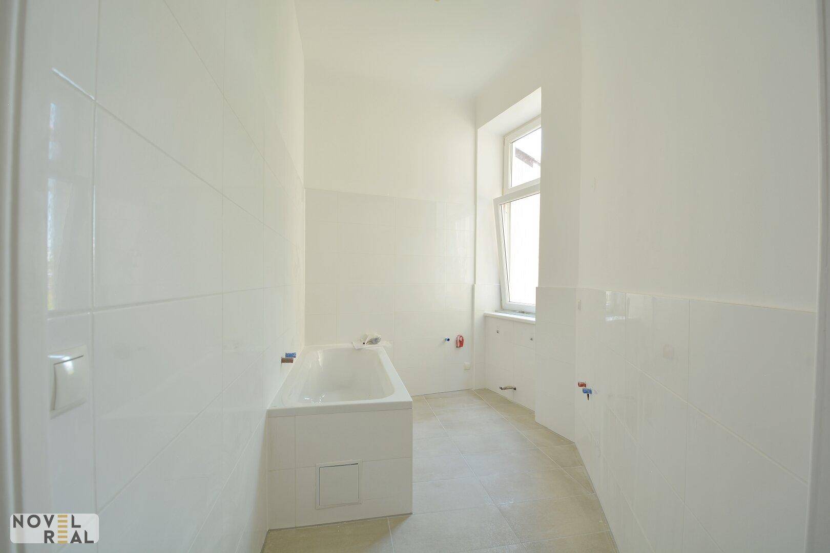 Unfertiges Badezimmer mit Badewanne, weißen Fliesen und einem Fenster für Tageslicht.