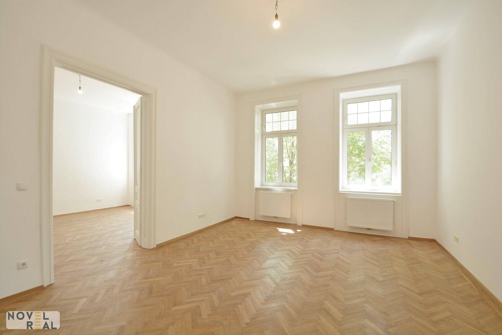 Leeres Zimmer mit Fischgrätparkett, weißen Wänden und großen Fenstern mit Blick ins Grüne.