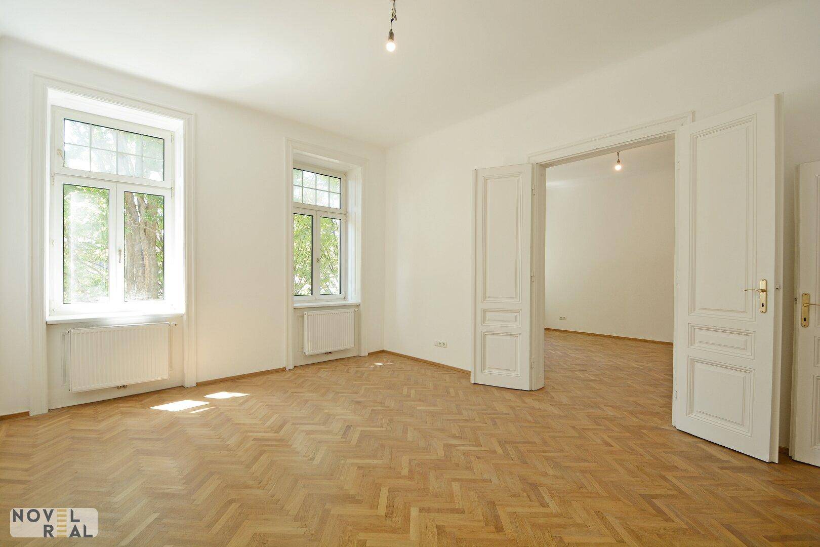Geräumiges, leeres Zimmer mit hellem Fischgrätparkett, hohen Decken und großen Fenstern.