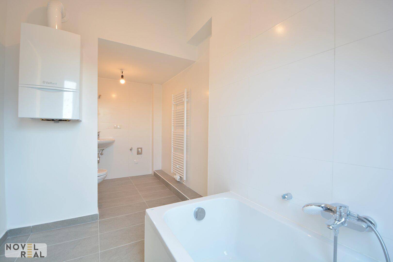 Helles Badezimmer mit Badewanne, weißen Fliesen, Handtuchheizkörper und Blick auf Waschbecken und WC.
