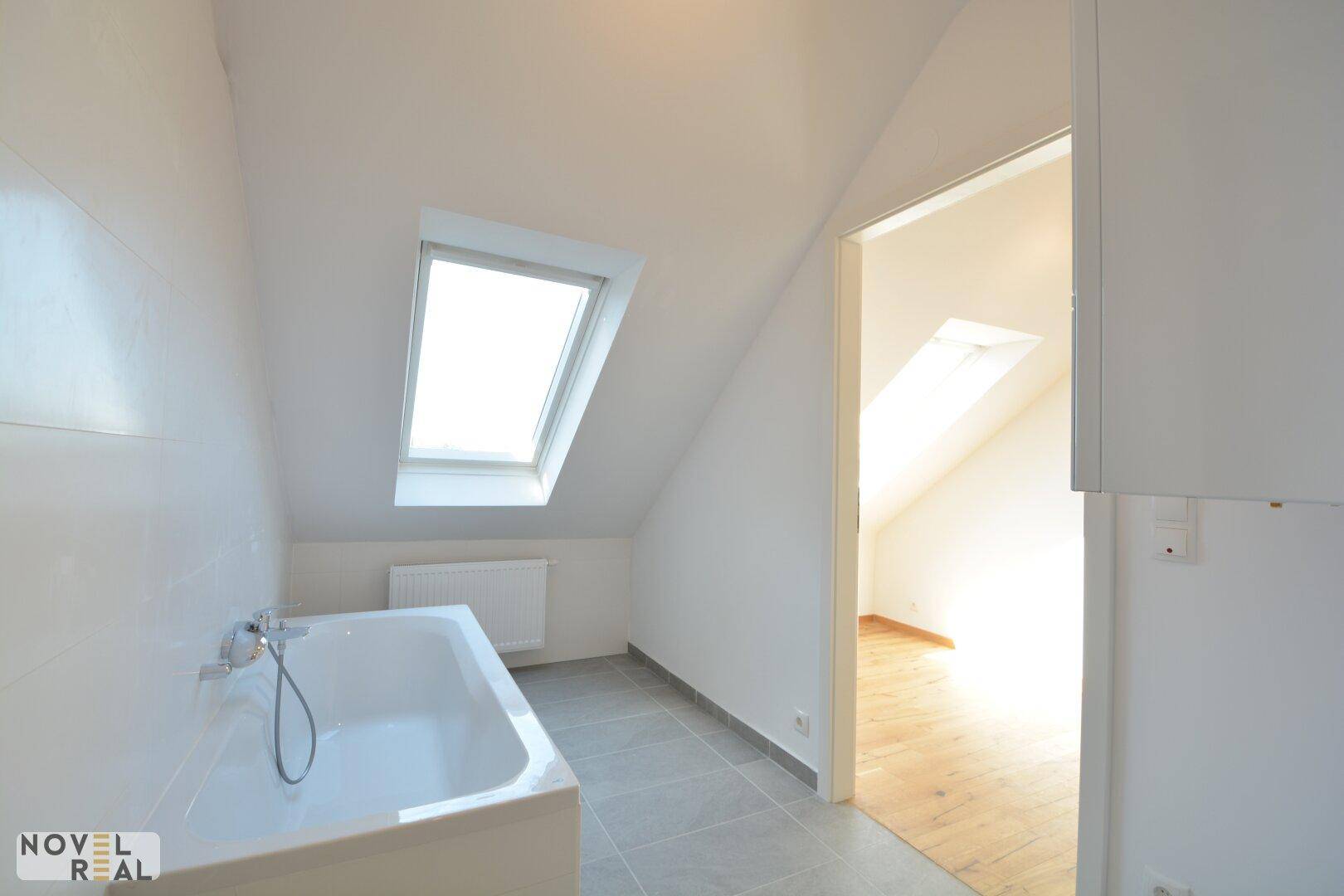 Modernes Badezimmer mit Badewanne, grauen Fliesen, Dachfenster und schrägen Wänden.