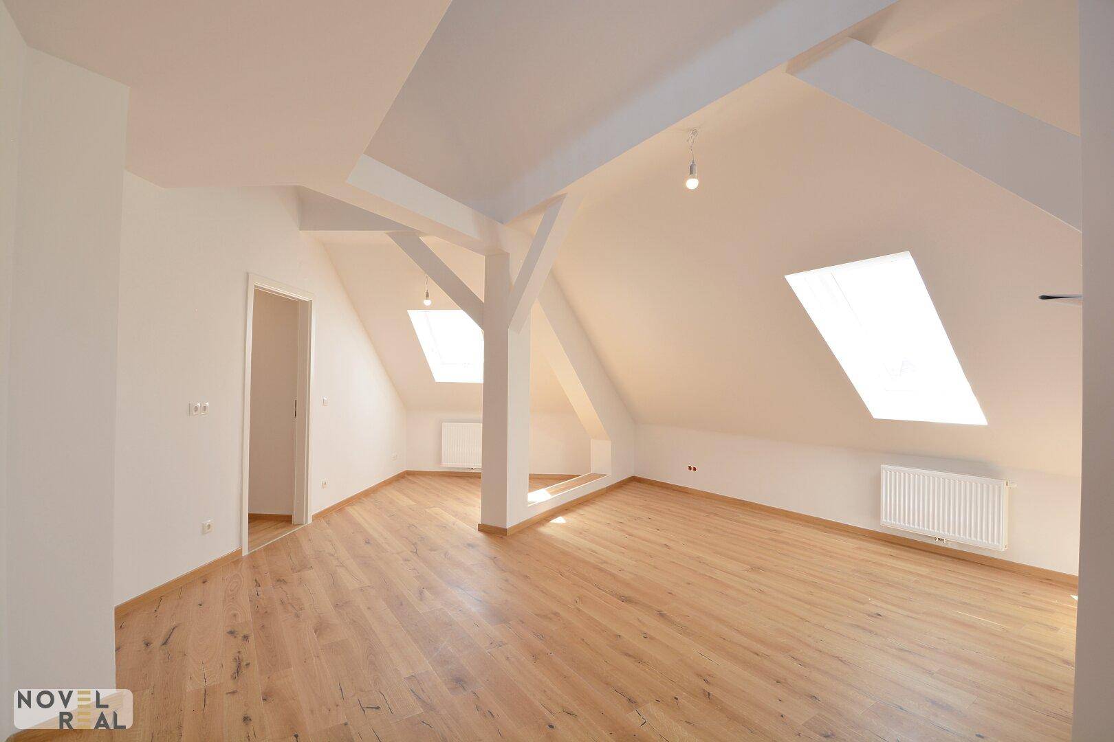 Heller, leerer Raum mit Dachschrägen, Holzboden und großen Dachfenstern, die viel Tageslicht hereinlassen.